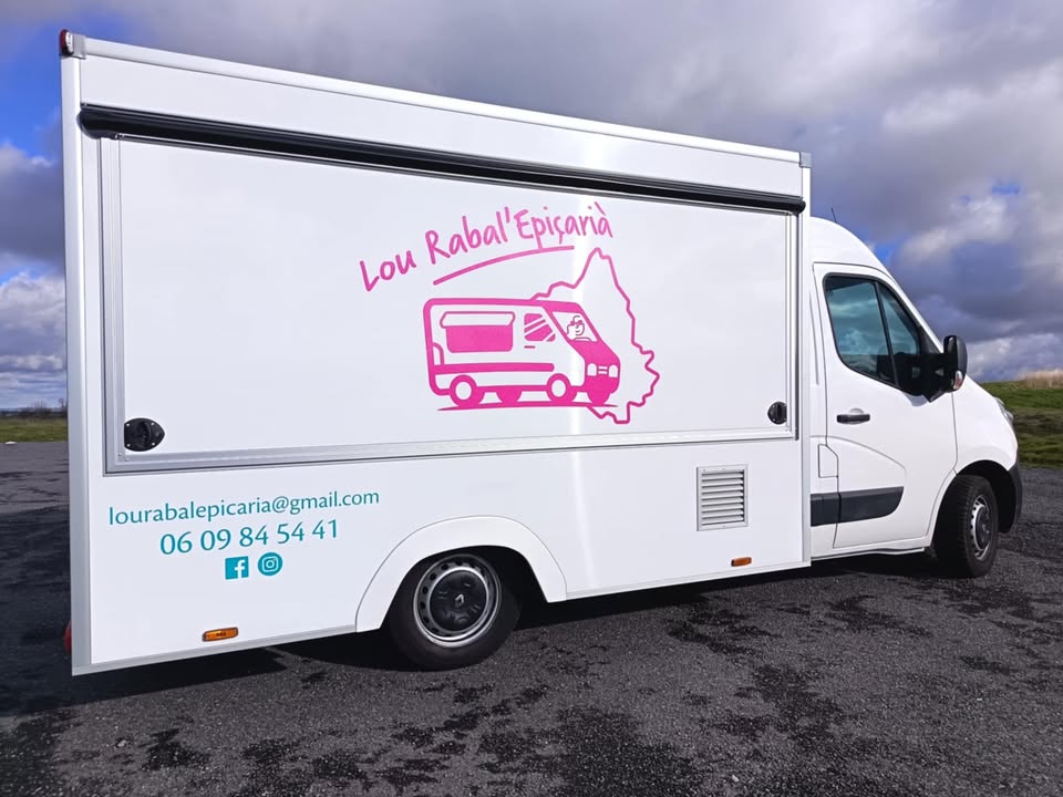 Camion-épicerie garé sur un sol bitumé. Sur le flanc latéral, une illustration rose fluo affiche le nom "Lou Rabal'Epicarià" et son logo.
