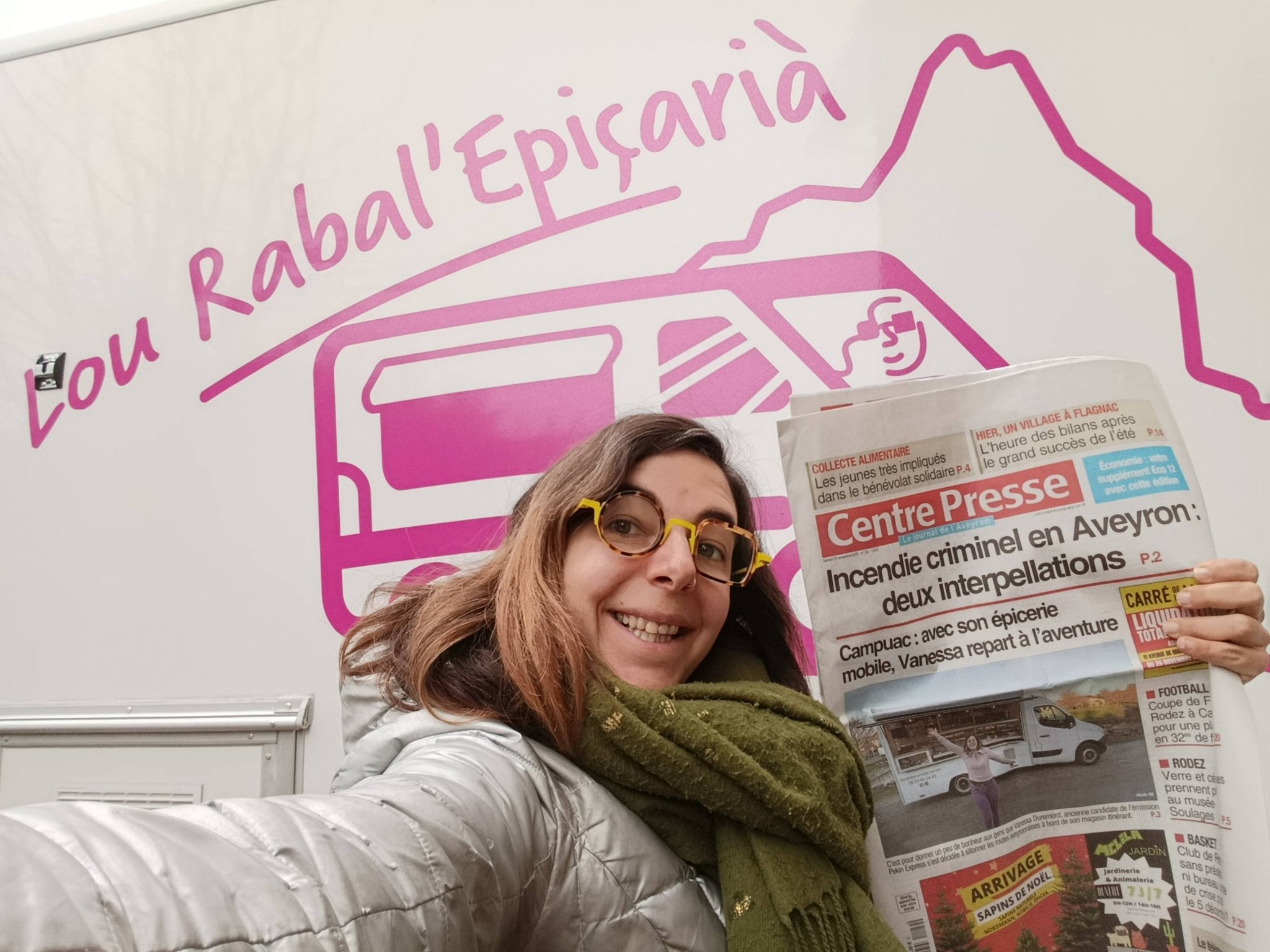 Vanessa, vêtue d'une veste argentée et d'une écharpe verte tenant une copie du journal Centre Presse se tient devant son camion-épicerie.